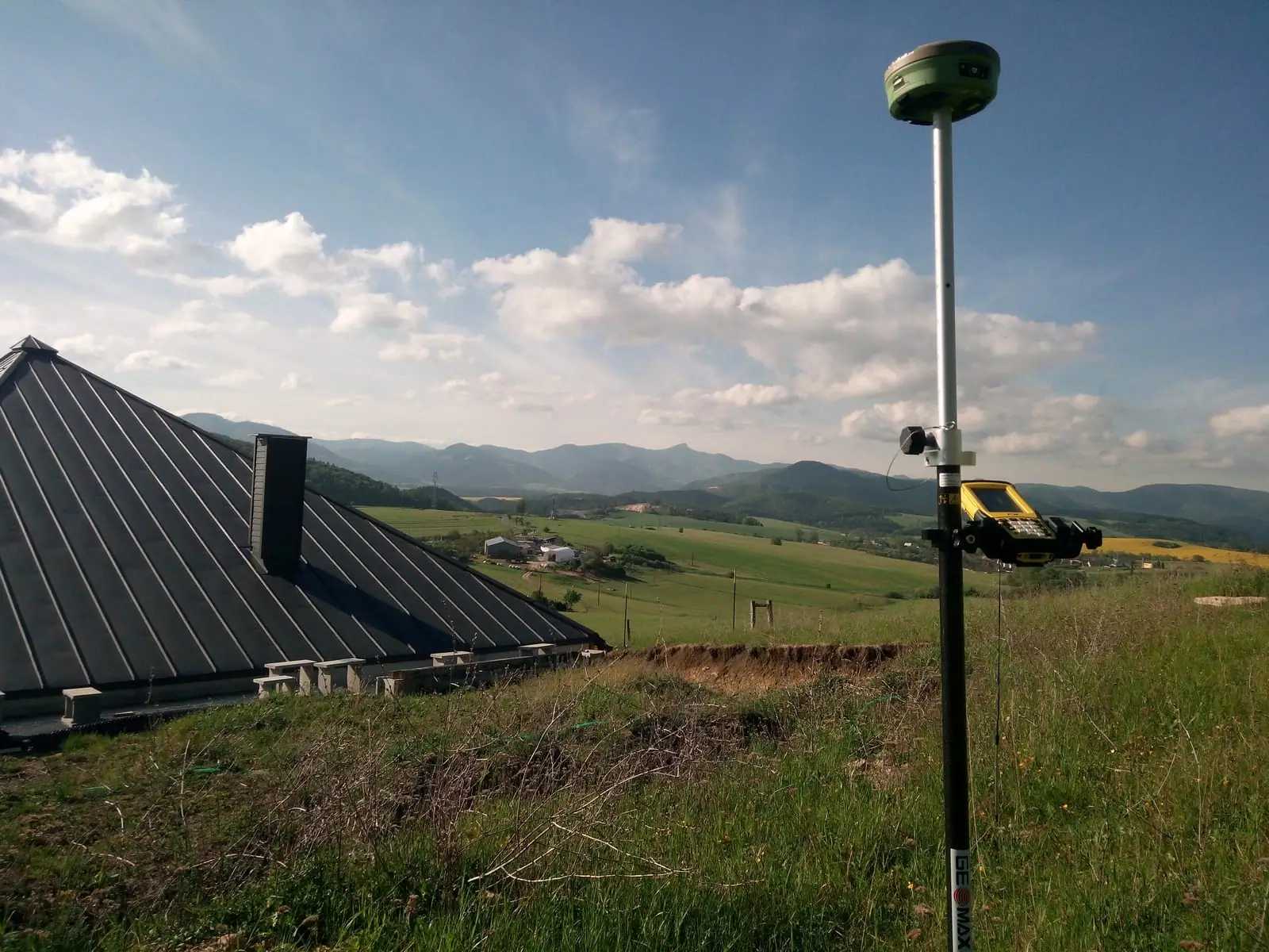 GNSS prijímač na výtyčke na svahu nad strechou domu s výhľadom do údolia