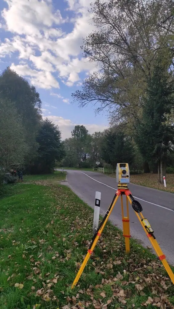 Statív s totálnou stanicou na tráve pri ceste lemovanej stromami